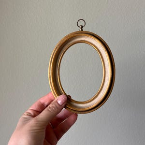 May include: Oval-shaped gold-toned picture frame with a cream-colored inner border. The frame has a decorative ring at the top for hanging. The frame is held by a hand.