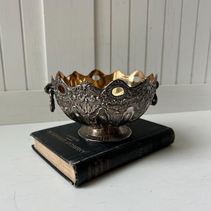 May include: Ornate silver bowl with a scalloped rim and gold interior, resting on a vintage book. The bowl features intricate floral and scrollwork designs, with small handles on either side. The book has a black cover with gold lettering.
