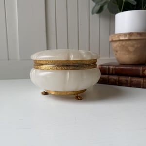 May include: A round, white, translucent stone trinket box with a fluted design. The container features a decorative gold-toned metal band around the center and rests on three small, ornate gold-toned metal feet. The lid is also fluted.