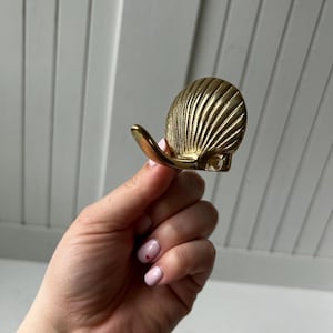 May include: A gold-colored seashell-shaped hook. The hook has a curved design and is made of metal. The seashell has detailed ridges and a smooth, polished finish. The hook is designed to be mounted on a wall or other surface.