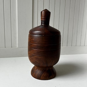 May include: A dark brown wooden jar with a lid. The jar has a rounded base and a cylindrical body with horizontal grooves. The lid has a conical top. The wood grain is visible, adding texture to the piece. The jar is likely used for storage.