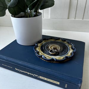 May include: A ceramic ring dish with a dark blue, tan, and black spiral design. Two rings rest in the center of the dish. The dish sits on a blue book next to a white potted plant with green leaves.