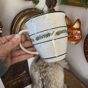 May include: A speckled white ceramic mug with a cream-colored handle. The mug features a green and black pinecone and leaf design, with two horizontal black lines. The rim is also trimmed in black. The mug is held by a person.