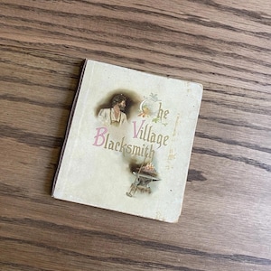 Puede incluir: Un libro antiguo titulado "The Village Blacksmith" con una ilustración de un herrero y sus herramientas. La portada es de color blanquecino con letras rosas y verdes y una pequeña ilustración de un herrero trabajando.