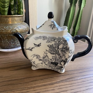 May include: Cream-coloured teapot with black accents. The teapot has a black handle, spout, and lid knob. The body is decorated with a black and white pastoral scene. A decorative item for the kitchen or dining room.