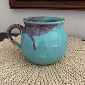 May include: A teal ceramic mug with a purple and white glaze. The mug has a rounded body and a handle.