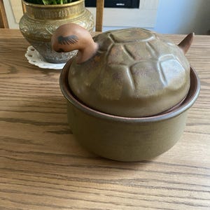 May include: A ceramic turtle-shaped container with a lid. The container is olive green with a brown turtle head and tail. The lid has a textured pattern. The container sits on a wooden table.