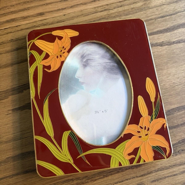 May include: A rectangular photo frame with a deep red enamel finish and gold-tone metal edges. The frame features an oval center opening and orange lily floral motifs with green stems. A paper insert displays the text 3½" x 5".
