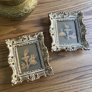 May include: Two rectangular picture frames with ornate, metallic gold-toned borders featuring intricate scrollwork and floral relief details. Each frame contains a print depicting ballet dancers in white tutus against a muted blue-grey background.