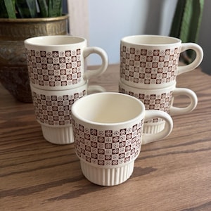 May include: A set of five cream-colored ceramic mugs with a brown checkered pattern featuring small floral motifs. Each mug has a rounded handle and a ribbed base design. The mugs are stacked in two columns with one mug placed in the front.