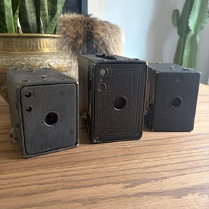 May include: Three vintage black box cameras arranged in a row. Each camera features a rectangular body with a central circular lens opening, a textured exterior finish, and small metal hardware components on the top and side panels.