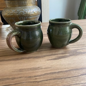 Könnte beinhalten: Zwei kleine, handgefertigte grüne Keramikbecher mit abgerundeten Körpern und gebogenen Griffen. Die Becher haben eine glänzende Oberfläche und stehen auf einer Holzoberfläche. Im Hintergrund befindet sich eine dekorative Messingvase.