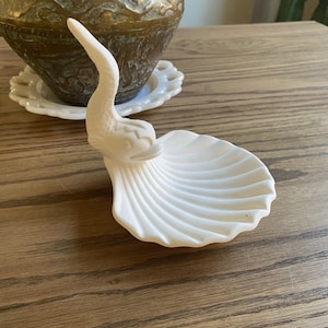 Puede incluir: Un plato blanco de cerámica con forma de cisne y base de concha. El cisne tiene un cuello largo y curvado y una cabeza detallada. El plato está sobre una superficie de madera.