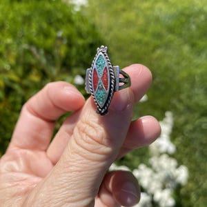 Puede incluir: Un anillo de plata con una pieza central en forma de marquesa incrustada con fragmentos de turquesa y coral. El diseño central está enmarcado por un borde de cuerda trenzada y la banda consta de tres hilos de plata paralelos a cada lado.