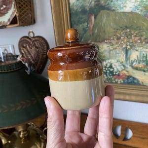 May include: A small, vintage ceramic jar with a lid. The jar is a light beige color on the bottom, transitioning to a tan band, and topped with a glossy brown glaze. The lid has a small, rounded knob. The jar is held in a person's hand.