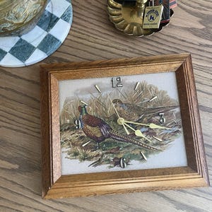 May include: A rectangular wooden framed clock with a pheasant illustration. The clock face features a detailed painting of two pheasants in a grassy field. The clock has gold-colored hands and numerals at 12, 3, 6, and 9.