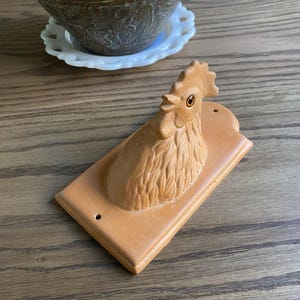 May include: A tan-colored rooster wall decor piece. The rooster is mounted on a rectangular wooden plaque. The rooster has a detailed comb, wattle, and feathers. Two small holes are drilled into the plaque for hanging. The decor is likely vintage.