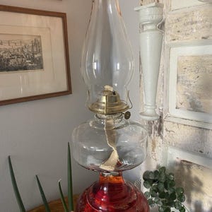 Peut inclure: Une lampe à huile composée d'une cheminée en verre transparent avec un sommet teinté ambre, d'un brûleur en laiton et d'un réservoir en verre transparent reposant sur une base en verre rouge profond ornée de motifs floraux gravés.