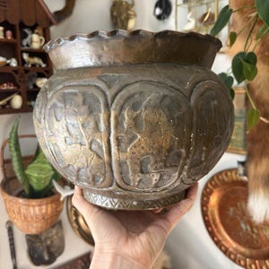 May include: A round, dark bronze-toned metal bowl with a scalloped rim. The exterior features embossed panels depicting figures and floral motifs, separated by vertical borders. The base is decorated with a repeating triangular pattern.