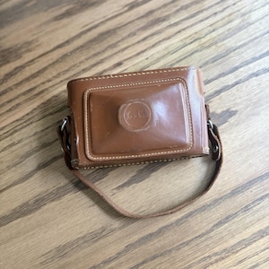 Peut inclure: Un étui pour appareil photo en cuir marron de forme rectangulaire avec une section centrale surélevée portant le logo Kodak en relief. L'étui comprend une sangle en cuir assortie fixée à des boucles latérales en métal pour protéger l'appareil.