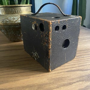 May include: A vintage black box camera with a textured leather-like exterior and a worn brown leather handle on top. The camera features a circular lens opening, two small viewfinder windows, a silver metal shutter button, and a metal winding key.