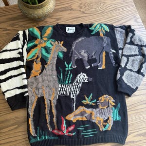 Peut inclure: Un pull en tricot noir à manches longues avec une scène d'animaux de safari colorée représentant une girafe, un éléphant, un zèbre et un lion. La manche gauche présente un motif rayé blanc et noir, tandis que la manche droite a un motif gris et noir rappelant la pierre. L'étiquette indique Joyce.