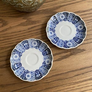 May include: Two round porcelain saucers with scalloped edges, featuring a white center and a blue and white patterned border consisting of alternating floral motifs and geometric diamond shapes arranged in a repeating circular design.