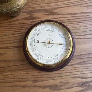 May include: A round analog barometer with a dark wood frame and a brass bezel. The white dial features black text including "Stormy," "Rain," "Change," "Fair," and "Very Dry," along with numerical scales. Two metal hands are centered on the face.
