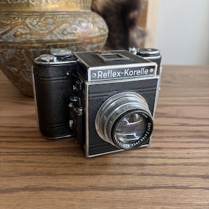 Könnte beinhalten: Eine Vintage-Kamera vom Typ Reflex-Korelle mit einem schwarz strukturierten Ledergehäuse und silbernen Metallakzenten. Die Vorderseite zeigt ein Carl Zeiss Jena Objektiv mit sichtbarem Text und ein silbernes Schild mit dem Markennamen Reflex-Korelle.