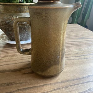 Może przedstawiać: Wysoki, brązowy ceramiczny dzbanek z uchwytem i pokrywką. Dzbanek ma dzióbek do nalewania i cylindryczny korpus. Ceramika ma nakrapiany, teksturowany wygląd. Dzbanek jest prawdopodobnie przeznaczony do serwowania napojów.