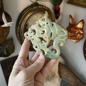 Puede incluir: Un colgante de jade verde pálido, tallado intrincadamente. El colgante presenta un diseño calado y arremolinado con un motivo de dragón o criatura mítica. El jade tiene un acabado liso y pulido, sostenido en una mano.