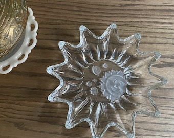 May include: A clear glass star-shaped dish with a frosted sun and moon design in the center. The dish has pointed edges and is sitting on a wooden surface. The dish is likely used for decorative purposes.