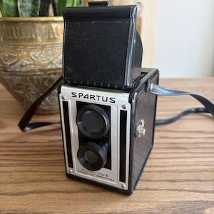 May include: A black and silver Spartus Full-Vue twin-lens reflex camera with a top-mounted viewfinder hood. The front face features the brand name Spartus and the model name Full-Vue Chicago, Ill. Made in U.S.A. printed in black text on a silver plate.