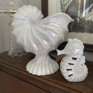 Może przedstawiać: Dwie iryzujące białe rzeźby ceramiczne. Jedna to duża muszla nautilusa, a druga to konik morski. Obie mają perłowe wykończenie. Rzeźby są wystawione na drewnianej powierzchni.
