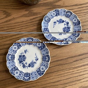 May include: Two small, round ceramic plates with scalloped edges. Each plate features a white center decorated with a blue floral wreath and a border composed of alternating panels of blue and white geometric patterns and stylized floral motifs.
