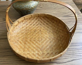 May include: A circular woven bamboo basket with a curved handle. The basket is light brown and features a tightly woven design. The handle is attached to the sides and arches upwards. The basket rests on a wooden surface.