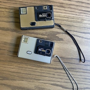 May include: Two rectangular Kodak Disc cameras with wrist straps. The top camera is black and gold with the text Kodak disc 3100. The bottom camera is silver and black with the text Kodak disc 4000. Both feature a flash, lens, and shutter button.