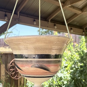 May include: A ceramic hanging planter with a wide, flared rim and a tapered base. The exterior features a beige base colour with horizontal bands in black and muted orange, accented by dark, curved brushstroke patterns encircling the body.