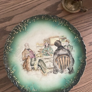 Puede incluir: Plato decorativo antiguo con una escena pintada a mano de personas reunidas alrededor de una mesa. El plato presenta un borde verde y dorado y una ilustración detallada con colores apagados. El plato es probablemente vintage y añade un toque de historia a cualquier decoración.
