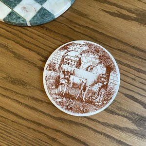 May include: A round, white ceramic coaster with a brown illustration of cows grazing near a stone structure. The coaster is on a wooden surface, with a marble and green checkered plate in the background.