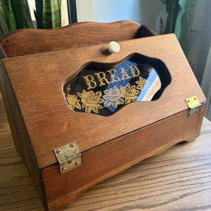 May include: A medium-brown wooden bread box with a hinged lid featuring a white knob. The lid has a central glass window with a scalloped edge, displaying the word BREAD in gold lettering above a gold floral rose pattern and brass hinges.