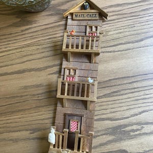 Op de afbeelding: Een houten wanddecoratie in de vorm van een gebouw met meerdere verdiepingen, met de tekst "MAIL CALL" op het dak. Het gebouw heeft balkons met kleine decoratieve items, waaronder een eend en een vogel. De kleurstelling is overwegend bruin en beige.