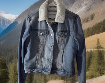 May include: A light blue denim jacket with a white, fluffy, sherpa-lined collar. The jacket has silver buttons and two chest pockets. The jacket is hanging on a white hanger.