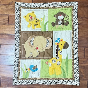 May include: A baby quilt featuring a patchwork design with various jungle animals. The quilt showcases a yellow tiger, a brown monkey, a tan elephant, a yellow giraffe, and a blue bird. The border has a brown and white spotted pattern.