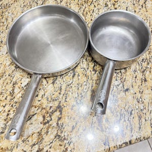 Può includere: Due padelle in acciaio inossidabile con manici lunghi. Una è una padella, l'altra un pentolino. Entrambe hanno una finitura metallica lucida e sono su una superficie marrone maculata. I manici hanno un foro per appenderle.