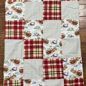 Puede incluir: Un edredón de retazos con cuadrados alternos de cuadros rojos y verdes, tela blanca con un patrón repetido de pequeños puntos blancos y tela blanca con una escena navideña de Papá Noel esquiando. El edredón es rectangular.