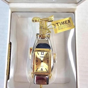 Może przedstawiać: Kolekcjonerski mini-zegar Timex w kształcie kija golfowego. Zegar ma złote wykończenie z prostokątną złotą tarczą, na której widnieją cyfry 12, 3, 6 i 9. Zegar jest w oryginalnym opakowaniu z metką Timex.