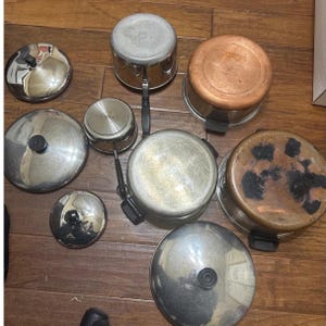 May include: A selection of stainless steel and copper pots and pans with lids. The pots vary in size and shape, some with black handles. The lids are stainless steel or have black knobs. Measurements are in cm.