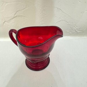 Könnte beinhalten: Eine rubinrote Glas-Sahnekännchen mit Henkel und Ausguss. Das Sahnekännchen hat einen runden Boden und einen geschwungenen Körper. Der Rand ist etwas dunkler als der Rest des Sahnekännchens. Das Sahnekännchen steht auf einer weißen Oberfläche.