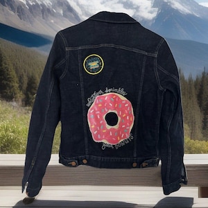 May include: Dark blue denim jacket with a pink donut graphic and the text "whatever sprinkle your donuts". The jacket also features a circular patch with text. A stylish and fun piece of apparel.